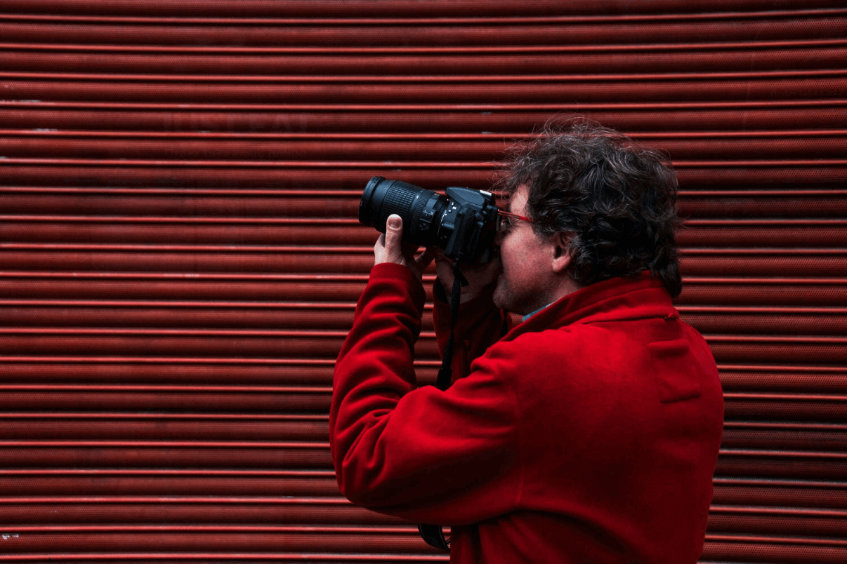 Einsteiger-Fotograf in roter Jacke hält eine DSLR-Kamera vor eine rote Metallwand